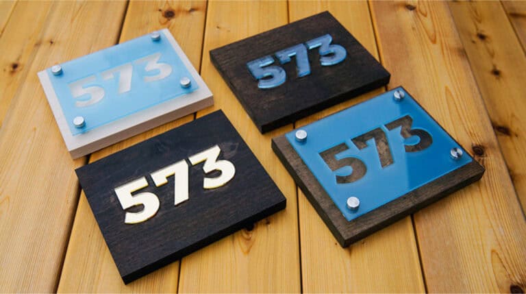 Quatre plaques en bois portant le numéro « 573 » dans différents styles et matériaux sont élégamment disposées sur une surface en bois, mettant en valeur des superpositions acryliques métalliques et bleues vibrantes.
