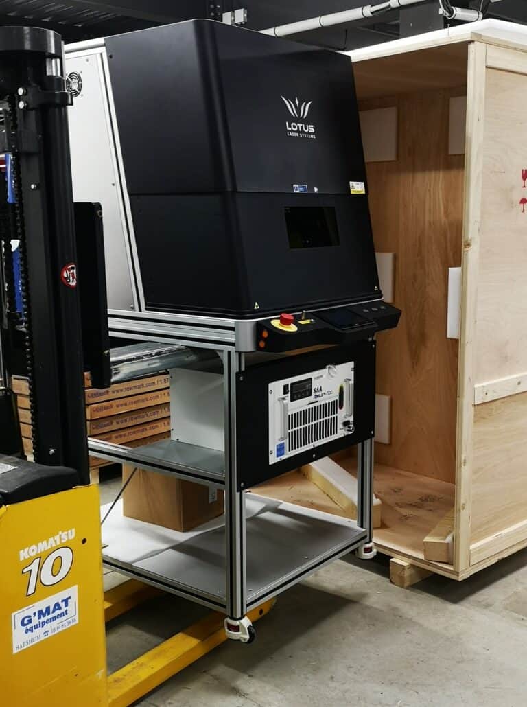 Une grande machine de gravure laser industrielle étiquetée « Lotus Laser Systems » repose sur un support métallique dans lentrepôt. À côté dune caisse en bois et perchée au sommet dune plate-forme mobile jaune Komatsu, cette impressionnante installation est équipée de la technologie de pointe Actu gamma tec.