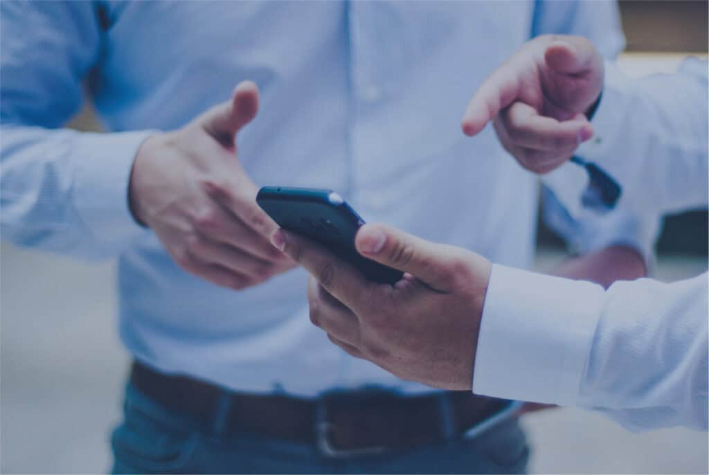 Deux personnes portant des chemises bleues discutent. L'une tient un smartphone et pointe l'écran pour proposer une technique d'assistance, tandis que l'autre fait un geste du pouce levé. L'accent est mis sur leurs mains et leurs bras.