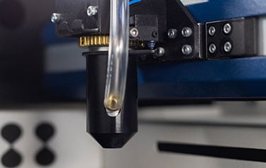 Gros plan dune buse dimprimante 3D en action, mettant en valeur ses composants noirs et métalliques avec précision. Un tube transparent relie la buse, encadrée par un boîtier bleu en arrière-plan. Cette configuration incarne la précision dune machine Epilog Fusion Maker, alliant art et technologie.