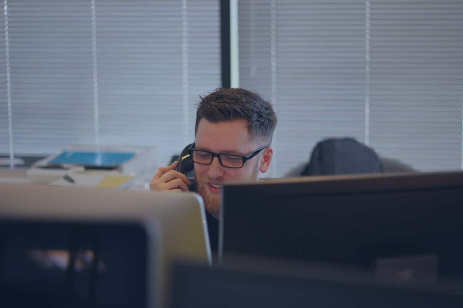 Un homme portant des lunettes est assis à un bureau, souriant tout en parlant au téléphone pour répondre à un client GAMMA-TEC. Des écrans d'ordinateur et du matériel de bureau l'entourent, tandis que des stores couvrent la fenêtre en arrière-plan.
