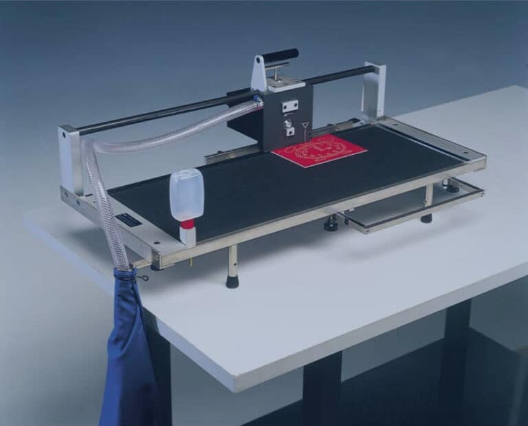 Une machine de sérigraphie textile sur une table, avec une surface plane sur laquelle est imprimé un imprimé rouge. Elle est équipée dun bras mécanique, de boutons réglables, dun tube transparent et dun petit récipient fixé. Le fond est un dégradé bleu uni.