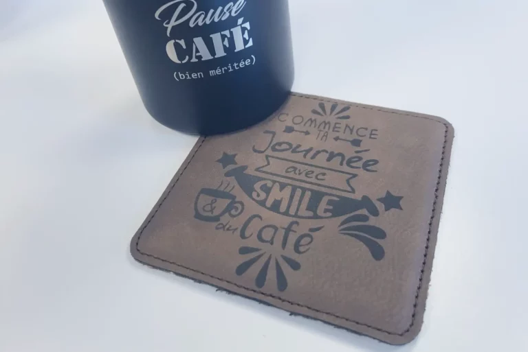Un dessous de verre marron avec la phrase « Commence ta journée avec smile et du café » à côté dune tasse noire qui dit « Pause café (bien méritée) » sur une surface blanche.