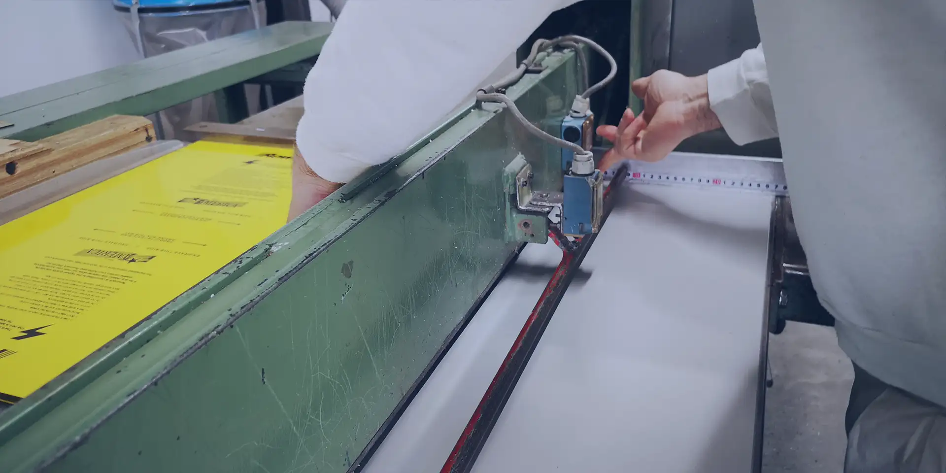 Une personne utilise une machine de sérigraphie et aligne une feuille de papier sur le plateau dimpression. La machine est peinte en vert et des feuilles jaunes avec du texte en arrière-plan. La personne porte une chemise blanche.
