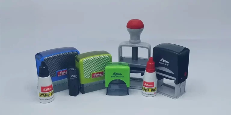 Une collection de différents tampons et bouteilles dencre est exposée. On y trouve des tampons auto-encreurs verts, bleus et noirs, ainsi que des bouteilles de recharge dencre rouge et blanche et un tampon en caoutchouc traditionnel avec une poignée rouge.