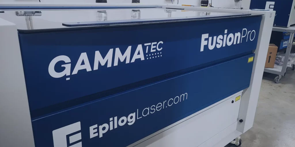 Gros plan dune machine laser Fusion Pro de Gamma Tec, présentant sa marque sur un panneau bleu dans un atelier. En ce qui concerne la technologie de pointe du secteur, le logo et le site Web epiloglaser.com sont bien visibles.