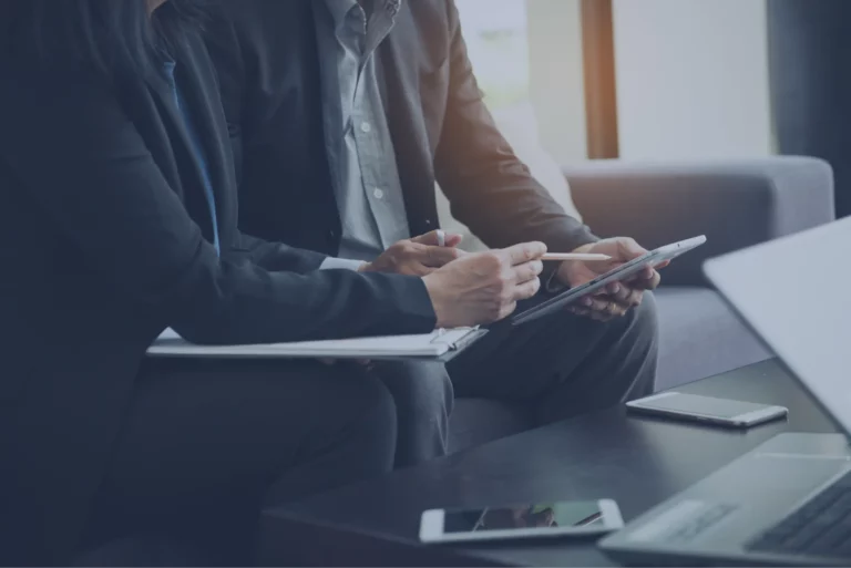 Deux personnes en tenue professionnelle sont assises sur un canapé et discutent autour dune tablette. Lune tient un stylo, tandis que des papiers et un smartphone sont posés sur la table. Un ordinateur portable partiellement visible au premier plan suggère un environnement de travail pour un revendeur de machines et de matières à graver.