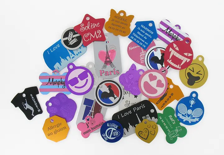 Une variété de plaques d'identification colorées pour animaux de compagnie, fabriquées avec précision à l'aide de machines laser et représentant des cœurs, des empreintes de pattes, des os et des motifs de tour Eiffel, sont affichées sur un fond blanc.