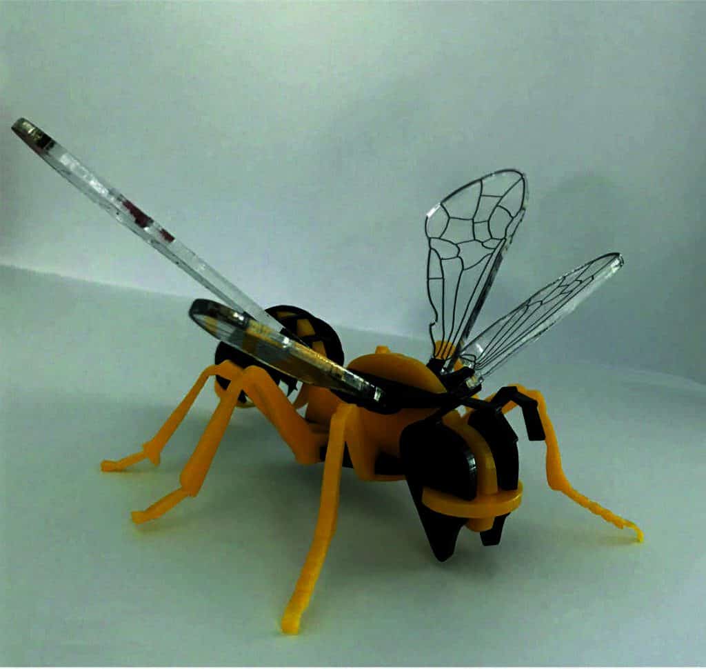 Une abeille robotique jaune et noire avec des ailes transparentes au design complexe se dresse sur une surface blanche, présentant une combinaison de caractéristiques mécaniques et dinsectes, ses composants gravés et découpés au laser pour plus de précision.