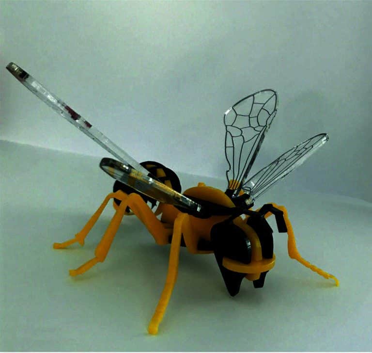 Une abeille robotique jaune et noire avec des ailes transparentes au design complexe se dresse sur une surface blanche, présentant une combinaison de caractéristiques mécaniques et dinsectes, ses composants gravés et découpés au laser pour plus de précision.