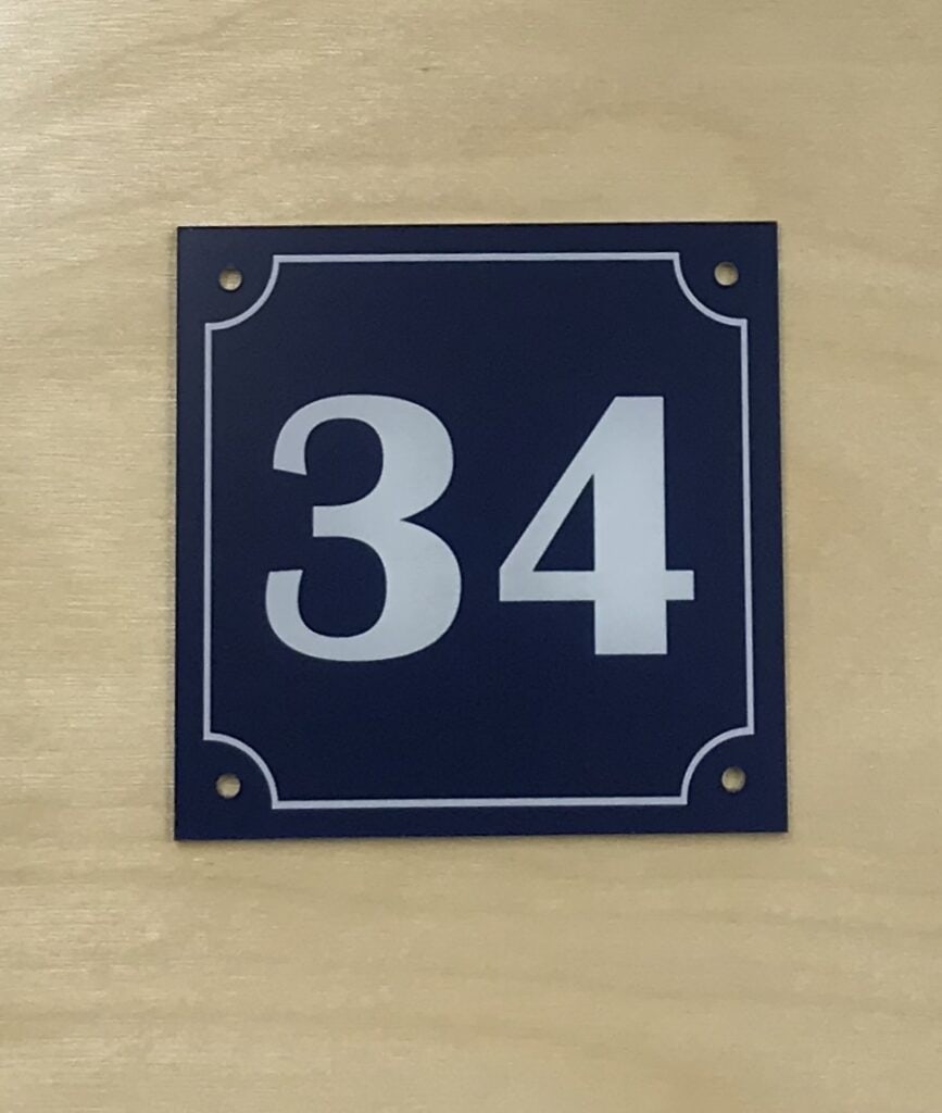 Un panneau rectangulaire bleu marine, fabriqué à l'aide d'un laser graveur, comporte des bordures blanches et le nombre 34 en caractères blancs gras, monté sur une surface en bois clair.
