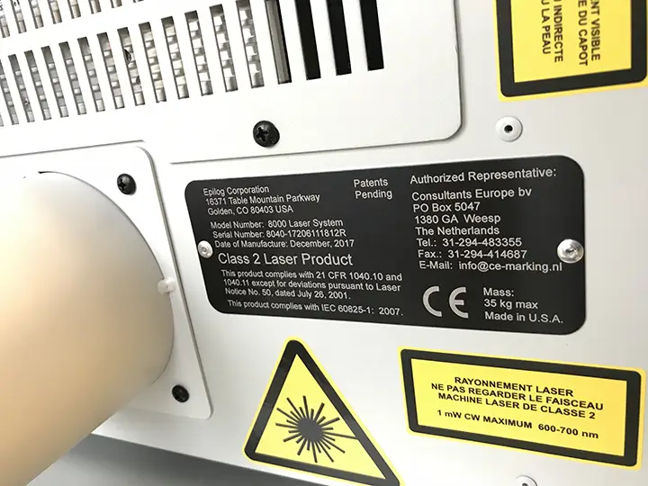Gros plan du panneau arrière d'une machine montrant plusieurs étiquettes d'avertissement et d'information, y compris des avertissements relatifs à la sécurité laser, les coordonnées du fabricant et du représentant, et les spécifications techniques. Un grand symbole jaune de danger laser est visible.