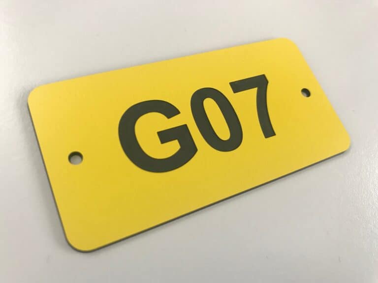 Une étiquette de machine rectangulaire jaune aux coins arrondis affiche le texte G07 en caractères noirs gras. Il y a deux trous, un de chaque côté de l'étiquette, probablement pour le montage. L'étiquette repose sur une surface claire.