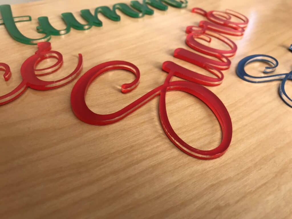 Gros plan sur des lettres acryliques cursives et colorées, notamment vertes, rouges et bleues, disposées sur une surface en bois clair. Les lettres brillantes, parfaites pour tout fournisseur de matière à graver, reflètent magnifiquement la lumière.