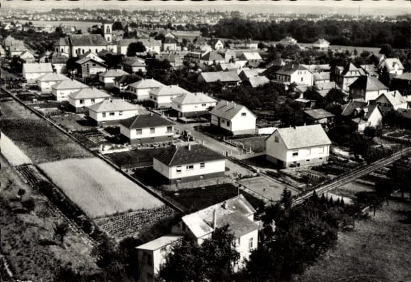 Vue aérienne dun quartier de banlieue avec rangées de maisons, jardins et arbres. Une église est visible en arrière-plan, à propos du mélange pittoresque de paysages résidentiels et ruraux entourés de vastes champs.