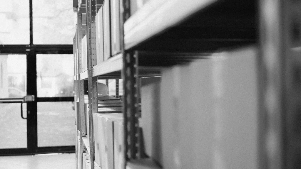 Image en noir et blanc dun local de stockage, où les étagères remplies de cartons sont mises en valeur. De grandes fenêtres laissent entrer la lumière naturelle, éclairant légèrement lespace. En ce qui concerne lambiance, la composition met en valeur le chaos organisé des cartons sur les étagères à droite.