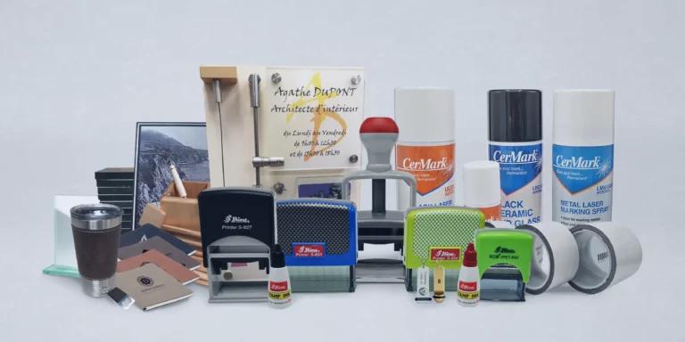 Une sélection de fournitures de bureau est présentée, notamment des tampons, des bombes aérosols pour produit de marquage, des porte-cartes de visite, des tubes adhésifs et un certificat dans un cadre transparent. Une image de montagne en arrière-plan ajoute une touche décorative.
