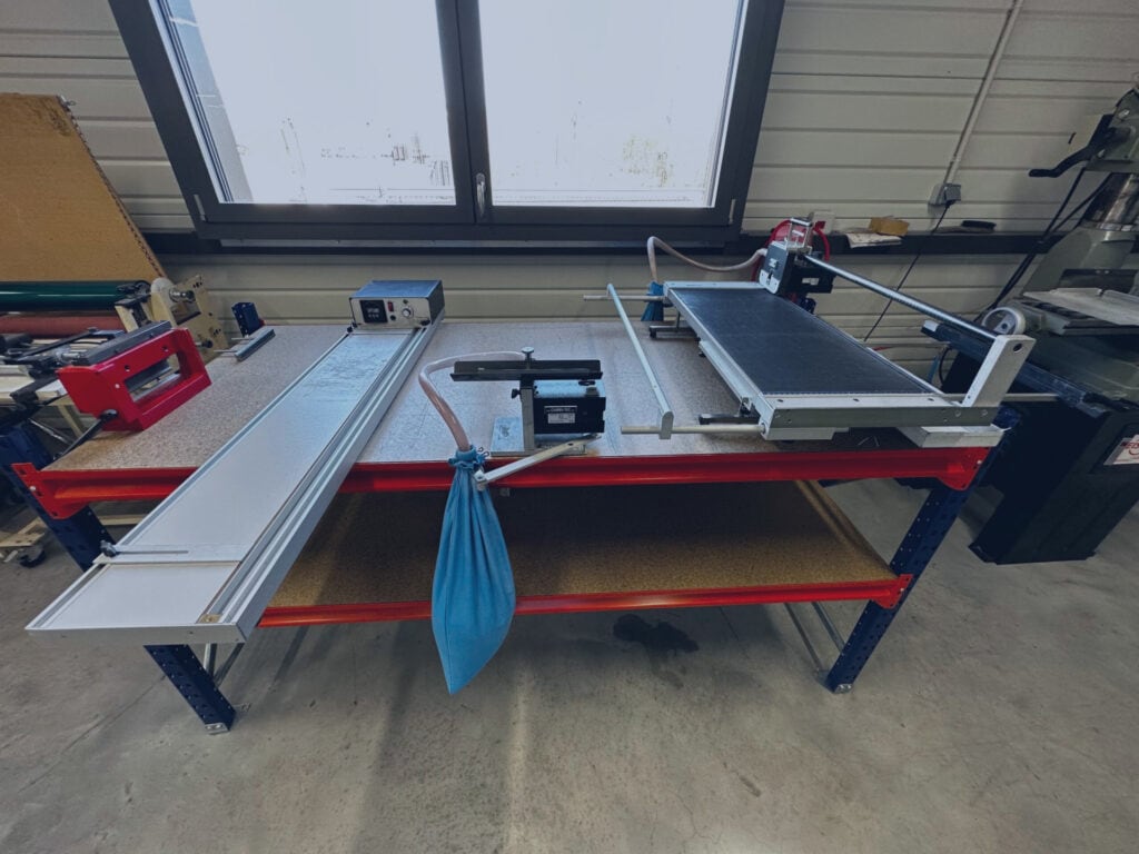Une table datelier dans un atelier lumineux présente diverses machines et outils, dont une scie à onglet, un long panneau blanc, un sac de collecte de poussière bleu et dautres équipements.