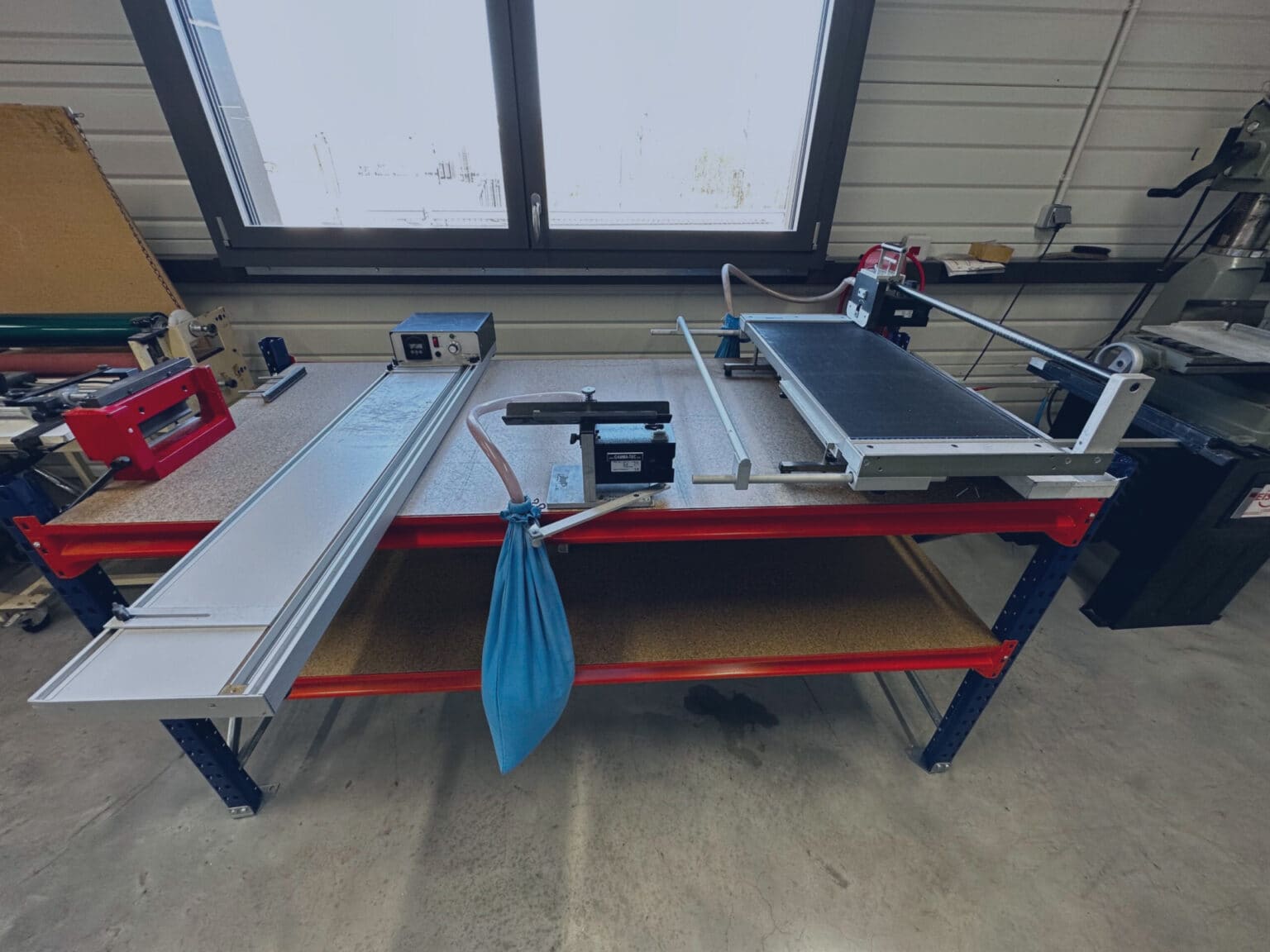 Une table datelier dans un atelier lumineux présente diverses machines et outils, dont une scie à onglet, un long panneau blanc, un sac de collecte de poussière bleu et dautres équipements.