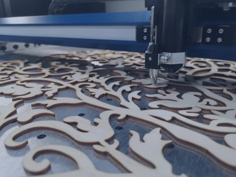 Une machine de découpe laser CO2 grave minutieusement un motif floral sur une feuille de bois. Le motif détaillé présente des tourbillons et des feuilles, témoignant dun savoir-faire précis. En arrière-plan, des parties de la machine et le cadre de support bleu sont visibles.