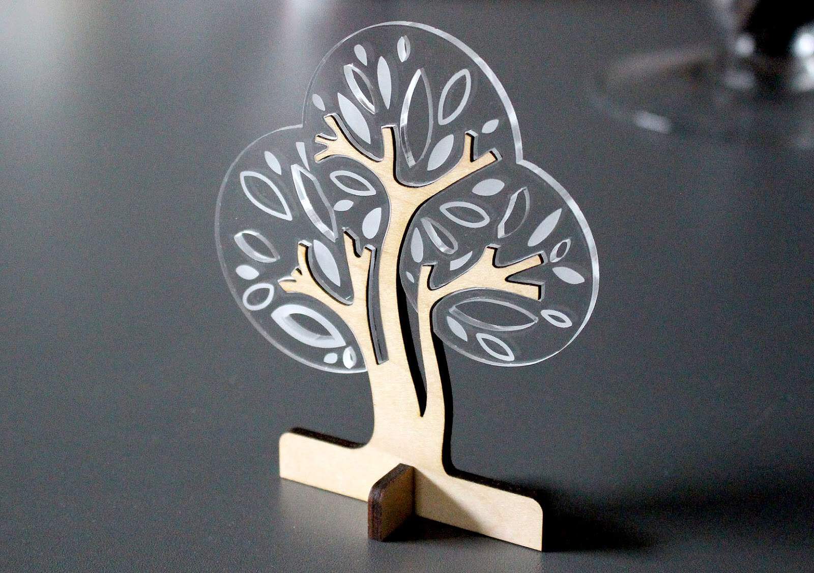 Une petite sculpture décorative darbre, gravée et découpée au laser, réalisée en bois et en plastique transparent présentant des motifs de feuilles, se dresse élégamment sur une surface grise lisse.