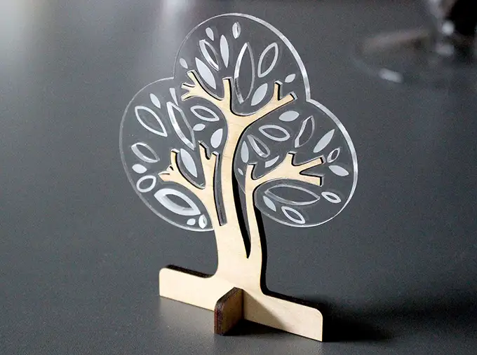 Une petite sculpture décorative darbre, gravée et découpée au laser, réalisée en bois et en plastique transparent présentant des motifs de feuilles, se dresse élégamment sur une surface grise lisse.