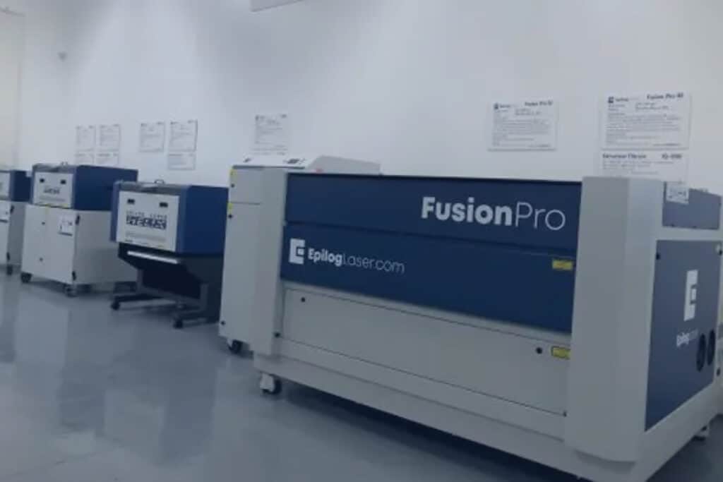 Dans un atelier moderne et bien éclairé, une rangée de machines de découpe laser Epilog Fusion Pro bleues et blanches est posée sur un sol brillant. Des instructions fixées au mur sont visibles en arrière-plan. Pour plus de détails ou pour planifier une démonstration, contactez GAMMA-TEC dès aujourdhui.