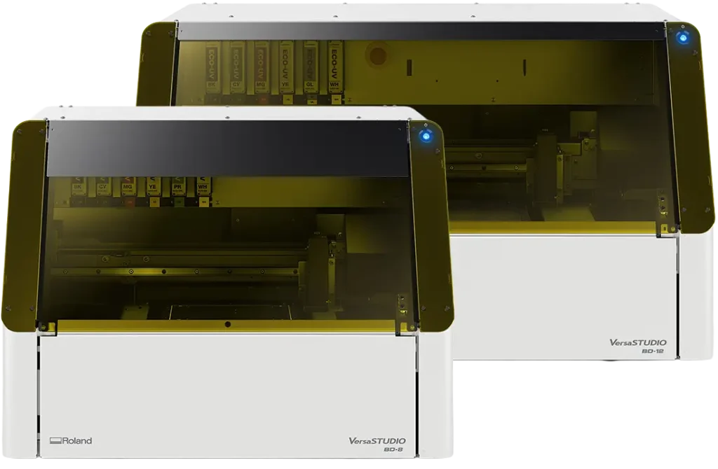 Deux appareils Roland VersaSTUDIO blancs, conçus pour l'impression d'objets, présentent des panneaux frontaux transparents teintés de jaune et sont placés côte à côte. Chaque machine porte le nom de son modèle dans le coin inférieur droit.