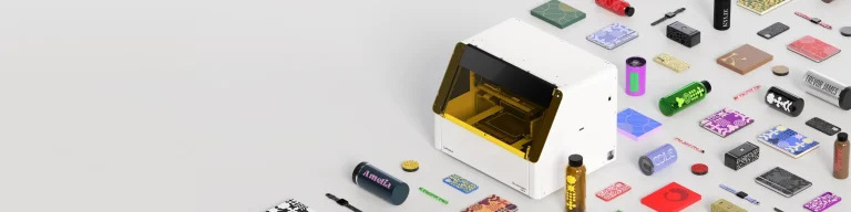 Une machine blanche avec une fenêtre teintée de jaune est posée sur une surface claire, entourée d'objets rectangulaires colorés, de tubes et de petits outils - tous bien rangés - laissant entrevoir un espace de travail impression objet en action.