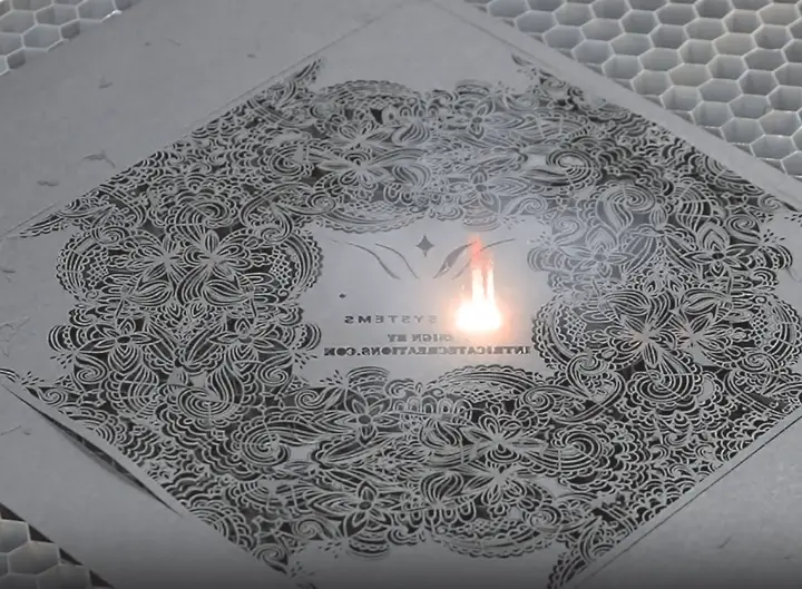 Un laser Marquage UV grave un motif floral symétrique complexe sur une pièce de métal, avec de la lumière visible et des reflets au centre du motif gravé. Le métal repose sur une grille en nid d'abeille.