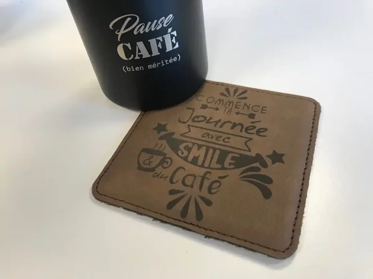 Une tasse noire avec linscription « Pause Café (bien méritée) » en texte blanc se trouve à côté dun sous-verre marron, orné dun texte français gravé au laser : « Commence ta journée avec un sourire & du café », rehaussé de graphismes décoratifs sur le thème du café.