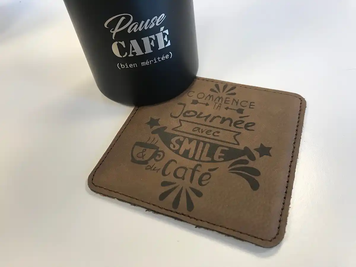 Une tasse noire avec linscription « Pause Café (bien méritée) » en texte blanc se trouve à côté dun sous-verre marron, orné dun texte français gravé au laser : « Commence ta journée avec un sourire & du café », rehaussé de graphismes décoratifs sur le thème du café.