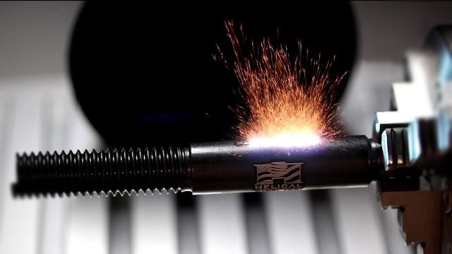 Des étincelles jaillissent lorsquune machine laser MOPA grave un objet métallique cylindrique étiqueté « Hélicoïdal ». Lobjet est solidement maintenu en place par un étau, larrière-plan flou soulignant laction et la précision de cette technologie de fibre avancée.