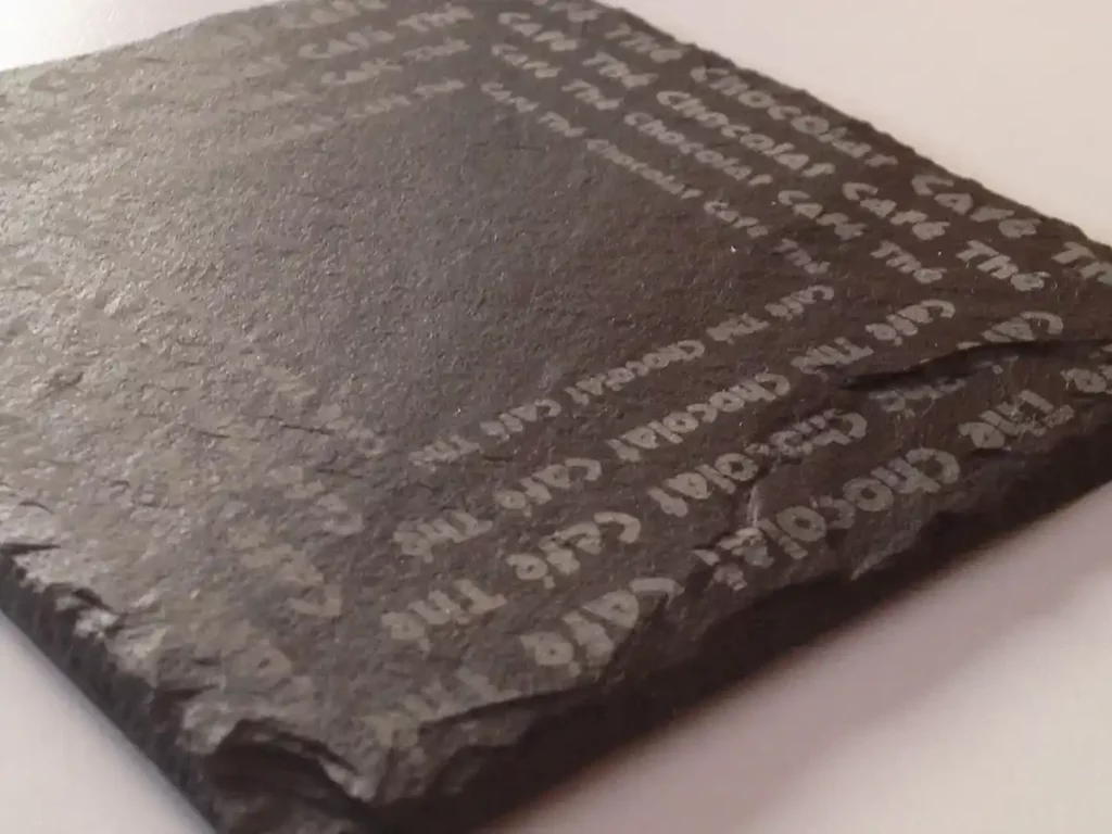 Un carreau dardoise texturé et foncé présente un texte blanc en relief. Ses bords rugueux et sa finition mate mettent en valeur les lettres en relief, créant un effet tactile superposé semblable à la gravure au laser sur ardoise sur un fond lisse.