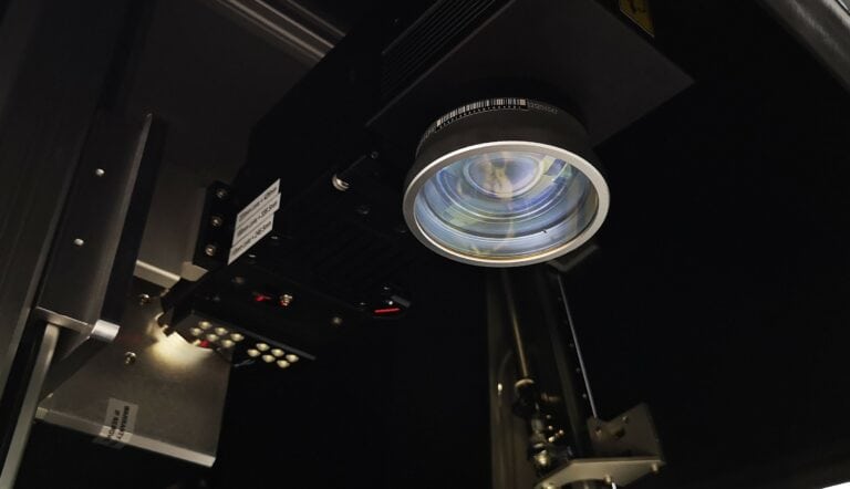 Gros plan sur un grand objectif dappareil photo ou un projecteur dans une pièce sombre, mettant en valeur les éléments en verre complexes. Les machines environnantes, avec leurs composants métalliques marqués au laser et leur technologie à fibre optique, créent une atmosphère industrielle et technique.