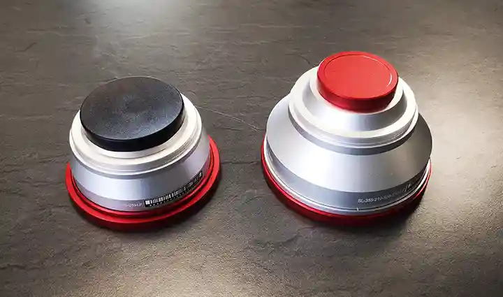 Deux objets cylindriques métalliques à sommet plat, ressemblant à des pièces de machines de précision, lun avec un sommet noir et lautre avec un sommet rouge, reposent sur une surface sombre. Tous deux ont un corps argenté et une base rouge, ce qui suggère leur rôle dans les procédés de gravure ou de laser.