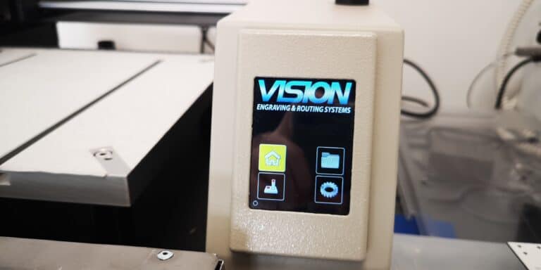 Gros plan du panneau de commande dune machine Vision Engraving & Routing Systems avec un écran éclairé affichant les icônes pour laccueil, le dossier et loutil. La petite machine de fraisage est dotée dun boîtier beige et léquipement de latelier est partiellement visible en arrière-plan.