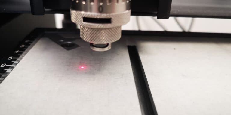 Gros plan dune découpeuse laser qui se concentre sur une feuille de métal avec son point laser rouge brillant. La surface présente une règle de guidage marquée sur la gauche, rappelant la précision nécessaire à la gravure sur verre à la machine. Lobjectif et la molette de réglage de la machine occupent le devant de la scène dans cette photo détaillée.