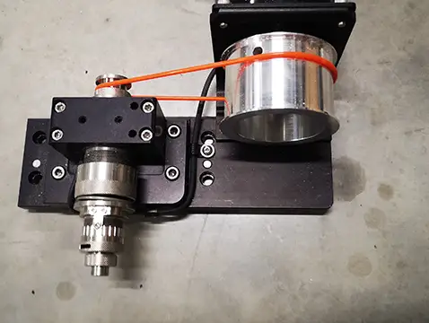 Dispositif mécanique posé sur un sol en béton, comportant à gauche un engrenage métallique relié par une courroie orange à un composant cylindrique brillant à droite. Lensemble est monté sur un socle noir avec des vis apparentes.
