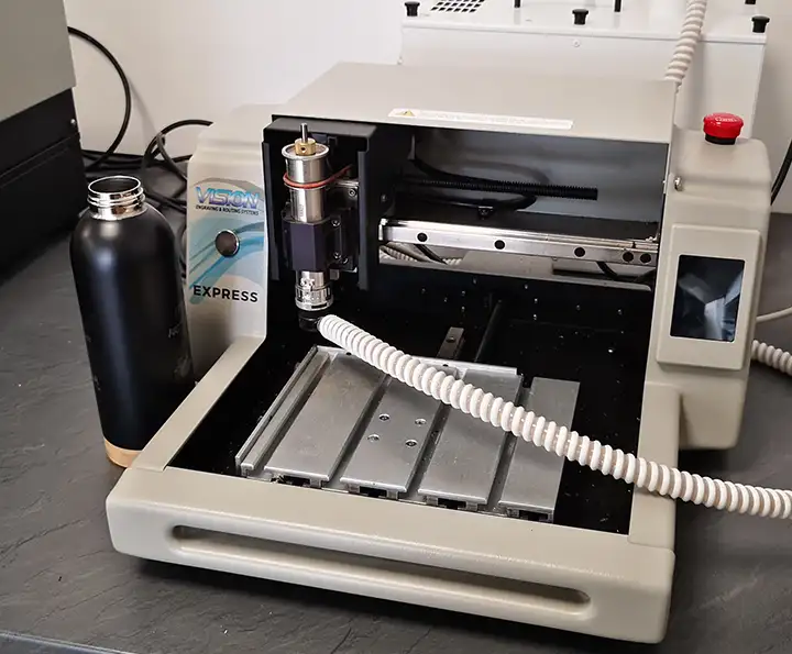 Une petite machine de fraisage est posée sur un plan de travail sombre, accompagnée dune bouteille deau noire. Cette fraiseuse CNC compacte est dotée dun petit lit de coupe et dun cordon dalimentation spiralé blanc branché dessus.