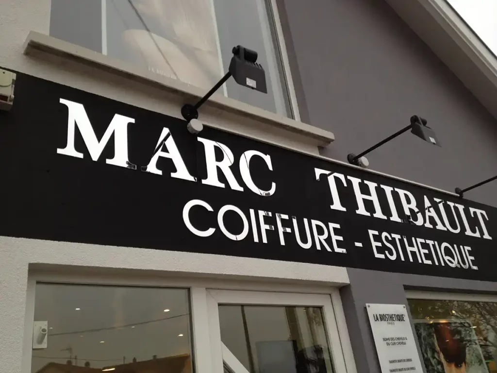 Lenseigne épurée « Marc Thibault Coiffure - Esthétique » orne lentrée du salon en blanc sur noir, avec deux lumières qui renforcent son allure. De grandes fenêtres offrent un aperçu de lintérieur, reflétant la précision des machines pour la signalisation.