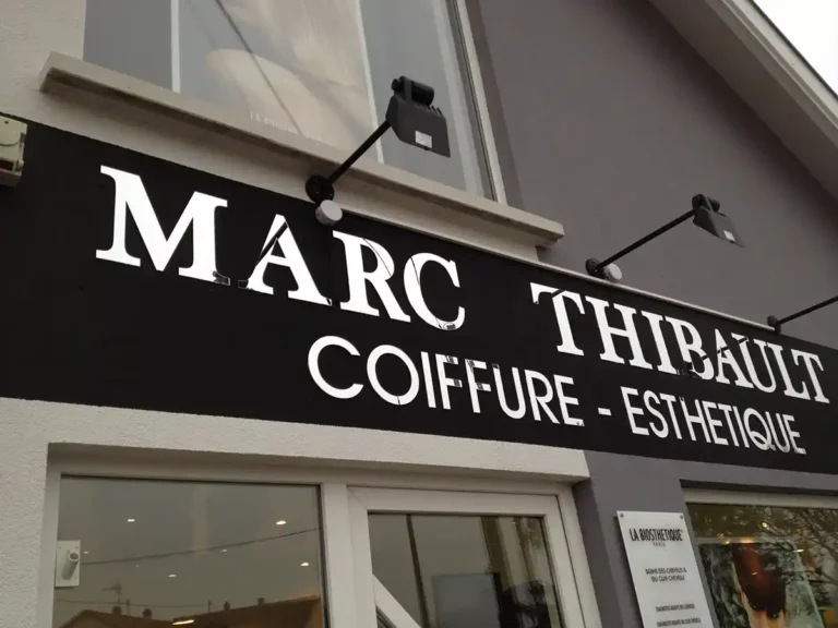 Lenseigne épurée « Marc Thibault Coiffure - Esthétique » orne lentrée du salon en blanc sur noir, avec deux lumières qui renforcent son allure. De grandes fenêtres offrent un aperçu de lintérieur, reflétant la précision des machines pour la signalisation.