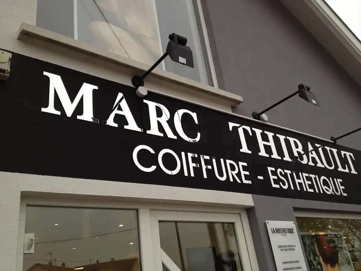 Lenseigne épurée « Marc Thibault Coiffure - Esthétique » orne lentrée du salon en blanc sur noir, avec deux lumières qui renforcent son allure. De grandes fenêtres offrent un aperçu de lintérieur, reflétant la précision des machines pour la signalisation.