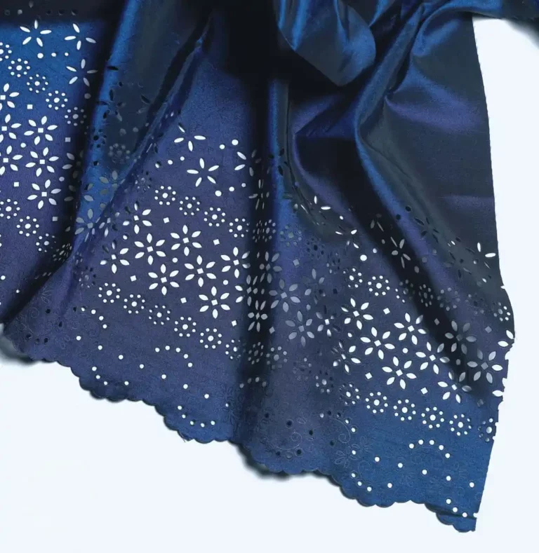 Un gros plan sur un tissu bleu foncé aux motifs floraux complexes et aux bords festonnés met en valeur une personnalisation textile exquise. Le motif présente de petites découpes délicates, créant une apparence de dentelle à la surface.