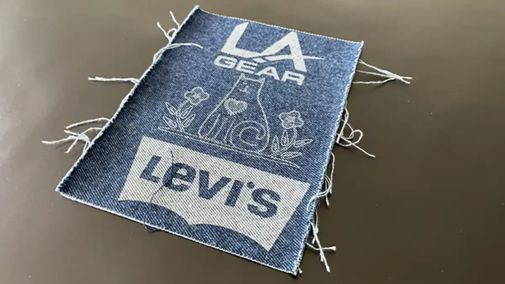 Un patch en denim parfait pour la personnalisation textile, avec « LA Gear » en haut, une forme géométrique avec des fleurs au centre et « Levis » en bas. Les bords effilochés ajoutent du caractère à sa surface unie.