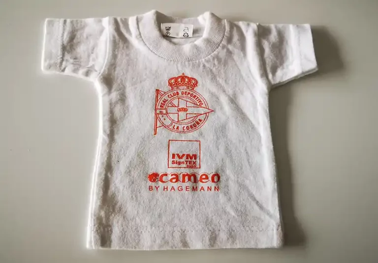 Un t-shirt blanc pour bébé avec le logo rouge du R.C. Deportivo La Coruña bien en évidence au centre, avec une couronne et une bande diagonale. En dessous, en texte rouge, on peut lire « IVM SignTex » et « cameo BY HAGEMANN », mettant en valeur lexpertise en matière de personnalisation textile.
