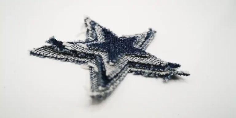 Un gros plan dun patch en denim effiloché en forme détoile, présentant une texture superposée et diverses nuances de bleu, incarne le charme du textile personnalisé. Le patch repose élégamment sur une surface blanche.