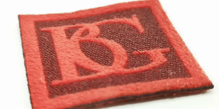 Gros plan dun patch texturé mettant en valeur les lettres rouges entrelacées « B » et « G » sur un fond carré rouge plus foncé, parfait pour les projets textiles de personnalisation.
