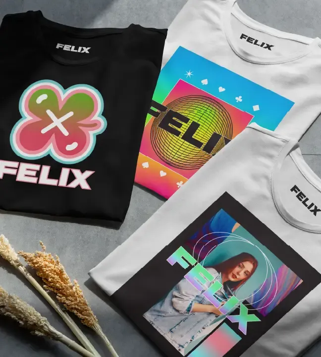 Trois t-shirts pliés de la marque FELIX sont disposés, chacun présentant des motifs audacieux rendus possibles grâce à l'imprimante/découpeuse jet d'encre. À proximité, des plantes séchées ajoutent une touche rustique à cette présentation accrocheuse.