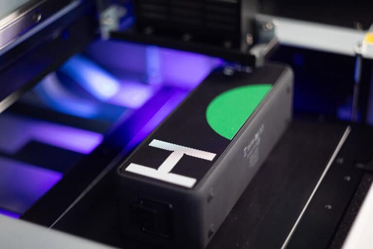 Une enceinte rectangulaire noire comportant un cercle vert et une lettre « H » blanche sur sa surface en maille fine est nichée à lintérieur dune machine avec un éclairage violet, créant un objet dimpression qui captive les sens.
